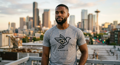 Confident Black man with low fade wearing Birdseye Core performance tee with eye-bird emblem and Seattle skyline at golden hour